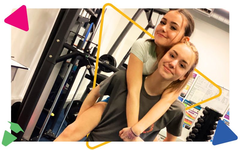 two smiling teenage girls in our gym. One of the girls is giving the other a piggy-back
